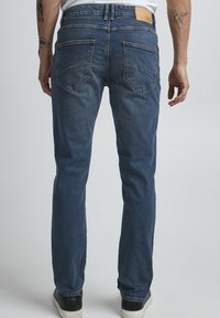Temno modri jeans prikazani s hrbtne strani, z zasnovo petih žepov, šivanimi detajli in usnjenim oznako na pasu.