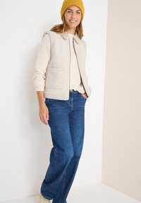 Gilet beige clair avec col et deux poches avant, superposé sur un pull crémeux. Associé à un jean bleu et à un bonnet en tricot jaune.