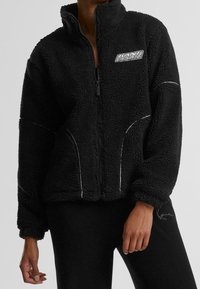 Chaqueta de forro polar negra con cuello alto, cremallera completa y detalles de costuras blancas. Presenta un parche con logo en el pecho y un corte relajado.