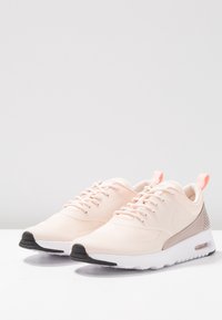 Light pink sneakers with a mesh upper, white midsole, and black outsole. Features include laces, a small branding detail, and a textured back panel.
