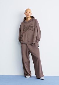 Brun hættebluse med grafisk print, der har snoretræk og frontlomme. Parret med løstsiddende brune sweatpants. Blød tekstur.