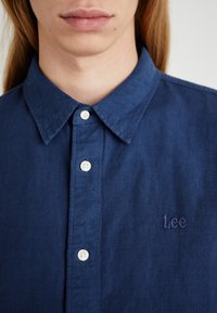 Niebieska koszula z guzikami w strukturze tkaniny, z kołnierzykiem w szpic i haftowanym logo "Lee", z białymi guzikami wzdłuż listwy.