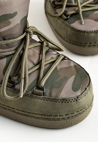Bottes à motif camouflage avec une tige en nylon vert texturé, semelle en caoutchouc renforcée et lacets tressés passés à travers des boucles en métal.