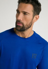 Royal blå atletisk t-shirt med rund halsudskæring og raglanærmer. Har et guld logo på venstre bryst. Glat stofstruktur.