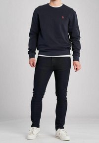 Marinblå sweatshirt med rund hals, ribbade muddar och en liten röd logotyp. Kombinerad med slim-fit mörka jeans och vita sneakers.