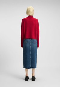 Personne avec des cheveux blonds courts portant un pull rouge, une jupe midi en denim bleu et des chaussures à plateforme noires, se tenant de dos sur un fond uni.