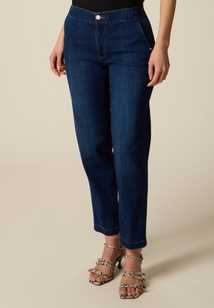 Donna che indossa jeans a gamba dritta blu scuro e sandali con tacco alto con stampa serpente e cinturini con fibbia, in piedi su uno sfondo semplice.