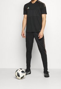 Svart träningsoutfit med kortärmad topp som har horisontella ränder och regnbågsliknande logotyp, ihopparad med svarta joggers och fotbollsskor. En fotboll ligger i närheten.