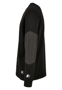 Sweat-shirt noir à manches longues avec coudières grises, poignets côtelés et deux petits patchs blancs avec logo près de l'ourlet et du poignet.