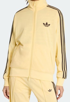 Persona que lleva un chándal Adidas amarillo claro con rayas negras en las mangas y los pantalones, y el logo de Adidas en el pecho y el muslo.