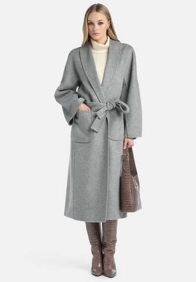 Lungo cappotto in lana grigia con cintura, maniche ampie e tasche frontali, indossato sopra un dolcevita crema. Gli accessori includono una borsa marrone con stampa cocco.