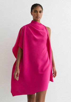 Kobieta w jasnoróżowej, asymetrycznej sukience bez rękawów z drapowanym materiałem na jednolitym białym tle.