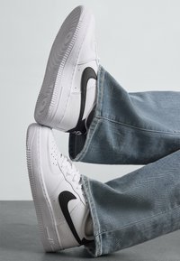 Biele tenisky Nike Air Force 1 s čiernym svužom, nosené s svetlom modrými džínami, soľe viditeľné na svetlo šedom pozadí.