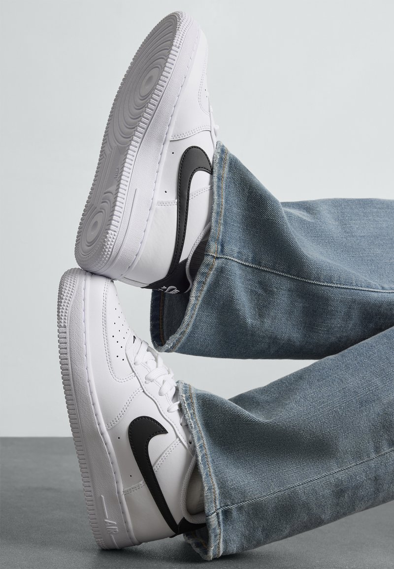 Biele tenisky Nike Air Force 1 s čiernym svužom, nosené s svetlom modrými džínami, soľe viditeľné na svetlo šedom pozadí.