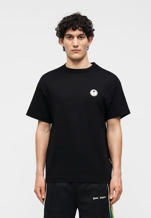 Νεαρός άνδρας με σγουρά μαλλιά φοράει μαύρο κοντομάνικο πουκάμισο με μικρό λογότυπο φοίνικα στο στήθος και μαύρο παντελόνι με λευκό κείμενο στον μηρό.