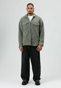 Green fleece jacket with front pockets, buttons, and a textured surface. Paired with loose black jeans and black shoes with pink laces.