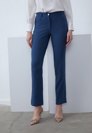 Femme portant un pantalon droit bleu marine, un chemisier blanc à manches longues et des talons hauts transparents avec des nœuds décoratifs.