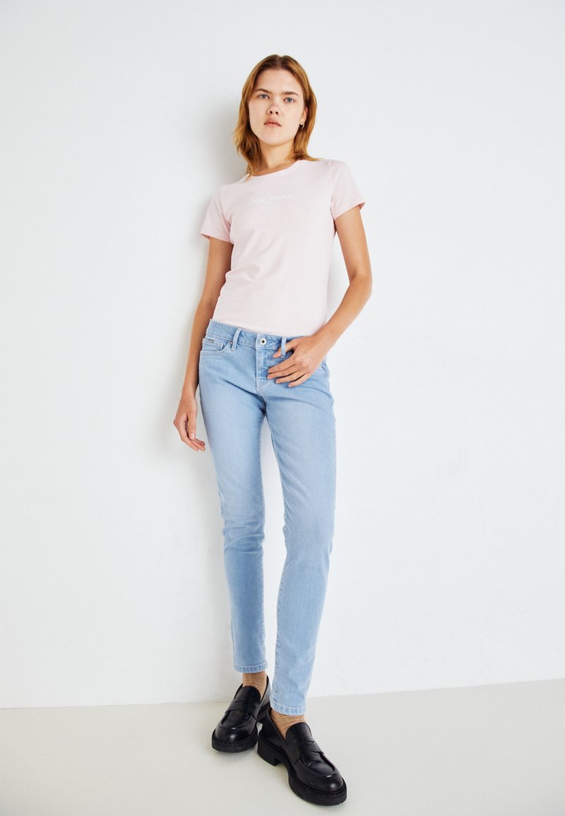 T-shirt rose clair à manches courtes avec du texte blanc, associé à un jean skinny bleu clair et des mocassins noirs. Design simple, style minimaliste.