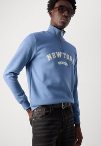 Světle modrá mikina na zip s nápisem "NEW YORK" v bílé a krémové barvě, nošena s tmavými džíny a černým opaskem. Model nosí sluneční brýle.