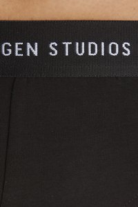 Sorte bomullstruser i svart med en bred elastisk linning som har "STUDIOS" i hvit strikket skrift. Glatt og tettsittende design.
