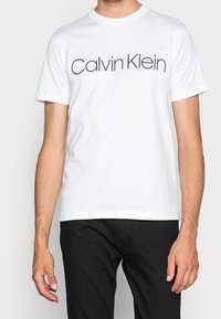 Bílý bavlněný tričko s kulatým výstřihem a krátkými rukávy, na přední straně s černým logem "Calvin Klein". Kombinováno s tmavými kalhotami.