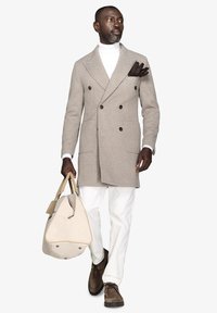 Lysbrun hørfilter dobbeltradet frakke, hvid turtleneck, hvide bukser, brune sko, holder en beige weekendtaske. Brun lommesquare detalje.