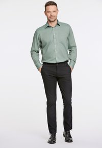 Camicia verde con bottoni e taschino al petto, abbinata a pantaloni neri e scarpe nere. Tessuto morbido, vestibilità su misura e design con colletto.