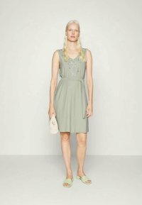 Vestido sem mangas verde claro com decote bordado, cintura cingida e silhueta fluida, acompanhado por sapatilhas verdes claras e uma pequena clutch.