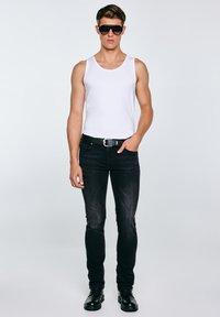 Fiatal férfi fehér ujjatlan pólóban, fekete farmerban, övvel, fekete csizmában és sötét napszemüvegben, egy egyszerű világos háttér előtt áll.