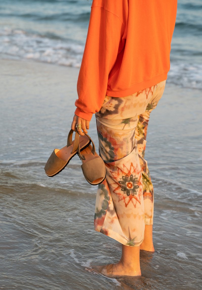 Des sabots en bois brun tenus dans une main, entourés de pantalons fluides à motif floral et d'un sweat-shirt orange, se tenant dans une eau peu profonde.