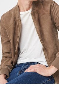 Man in een bruine suède jas over een wit T-shirt en blauwe jeans, zittend met één hand op zijn dij tegen een effen achtergrond.