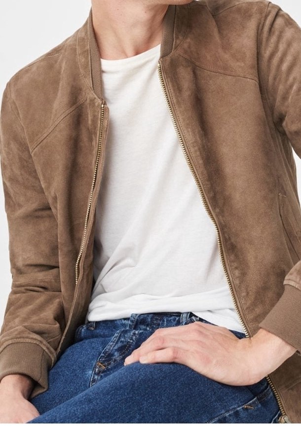 Man in een bruine suède jas over een wit T-shirt en blauwe jeans, zittend met één hand op zijn dij tegen een effen achtergrond.