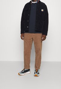 Giacca di velluto a coste nera sopra un polo blu navy, pantaloni di velluto a coste beige e sneakers grigie con accenti arancioni. Design minimalista e dettagli di texture.