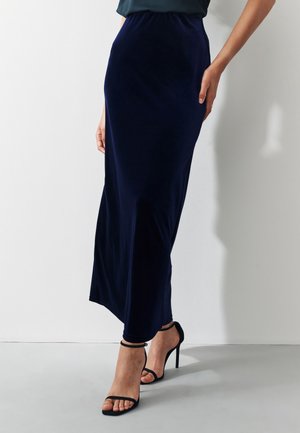 Femme portant une jupe en velours bleu marine longueur cheville et des sandales noires à talons hauts, debout contre un mur blanc uni.