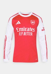 Rød fodboldtrøje med lange ærmer og hvide accenter, der har et struktureret mønster, logo på brystet og teksten "Emirates". Tre striber på skuldrene.