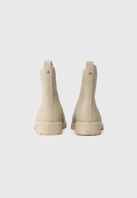Beige semskede ankelstøvler med flat gummisåle, elastiske sidepaneler og en metallaksontering bak, med et enkelt design.