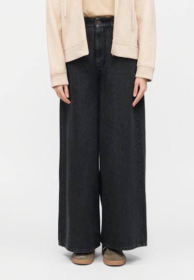 Personne portant un jean noir taille haute à jambes larges, une veste zippée beige, un haut beige et des baskets vert olive, debout devant un fond blanc.