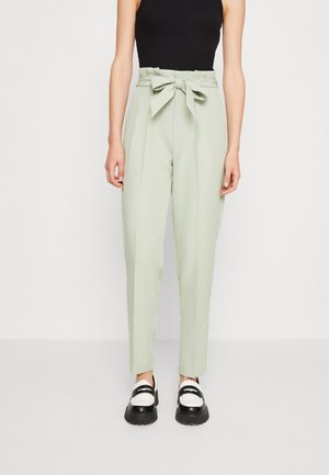 Femme portant un pantalon taille haute vert clair avec une ceinture nouée, un haut noir sans manches, et des mocassins épais noir et blanc, debout sur un fond blanc.