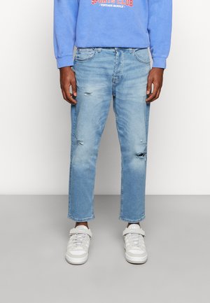 Person, die hellblaue, abgenutzte Jeans, weiße Turnschuhe und einen blauen Pullover trägt und vor einem einfarbigen Hintergrund steht.