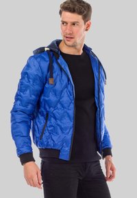 Bright blue quilted jacket with a black zip, fitted cuffs, and an attached hood. Features a textured surface and subtle logo detail.
