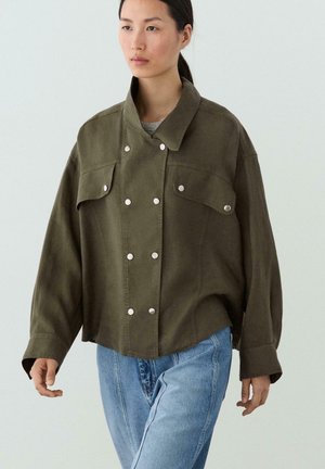 Femme aux cheveux foncés en queue de cheval, portant une veste ample vert olive avec des boutons argentés et un jean bleu, marchant devant un fond clair uni.