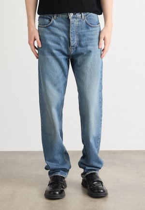 Person som har på seg løse lyseblå jeans og sorte lærloafers st�år på et grått gulv mot en enkel hvit bakgrunn.
