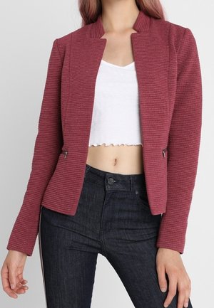 Vrouw draagt een geribbelde donkerroze blazer over een witte cropped top met zwarte high-waisted jeans, staand tegen een effen achtergrond.