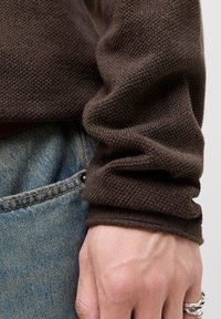 Hånden hviler ved siden, iført sølvfarvet, struktureret ring, brun strikket sweaterærme og lomme på blå denimjeans synlig.