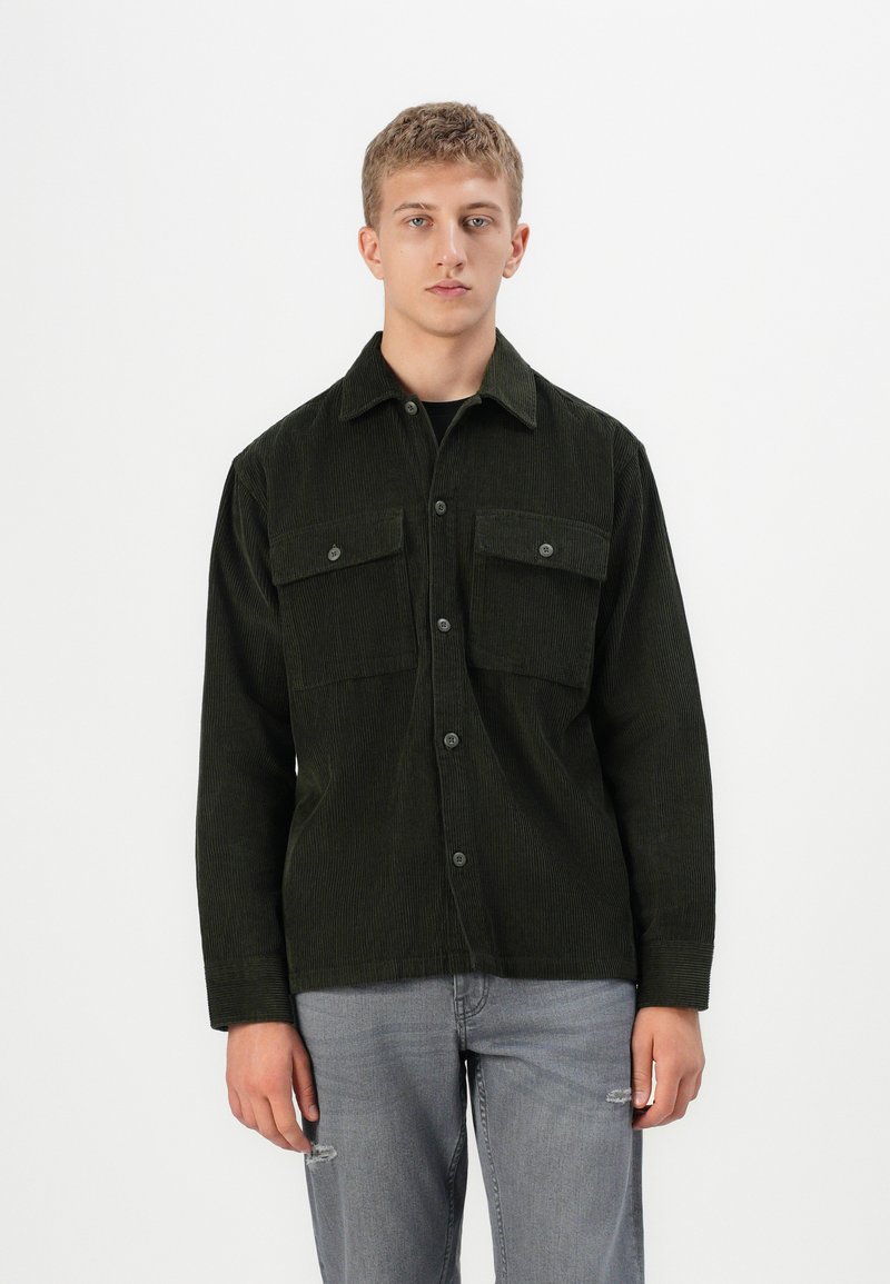 Un jeune homme portant une chemise boutonnée en velours côtelé vert foncé et un jean déchiré gris clair, se tenant contre un fond blanc uni.