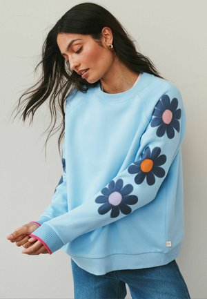 Žena s dlouhými tmavými vlasy, oblečená v světle modré mikině zdobené velkými tmavě modrými květy s barevnými středovými částmi na rukávech.