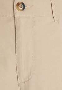 Nahaufnahme eines beigen Stoffes von einer Hose, mit sichtbarem Knopf, Taschennaht und glatter Oberfläche mit diagonalem Webmuster.