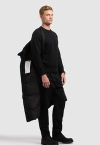 Homme vêtu entièrement de noir tient un manteau matelassé noir sur une épaule, sur un fond uni gris clair.