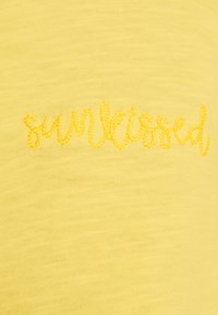 Tessuto giallo con una trama testurizzata e leggermente a coste. Presenta il testo ricamato "sunkissed" in una tonalità di giallo simile, creando un contrasto sottile.
