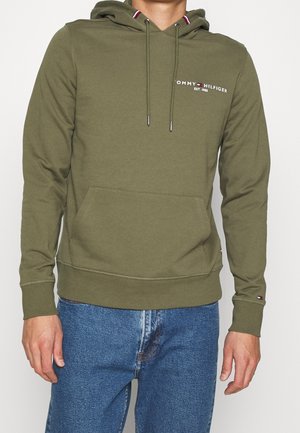 Muž ve světlounce zelené mikině Tommy Hilfiger s přední kapsou a modrých džínách, stojící před jednobarevným pozadím.